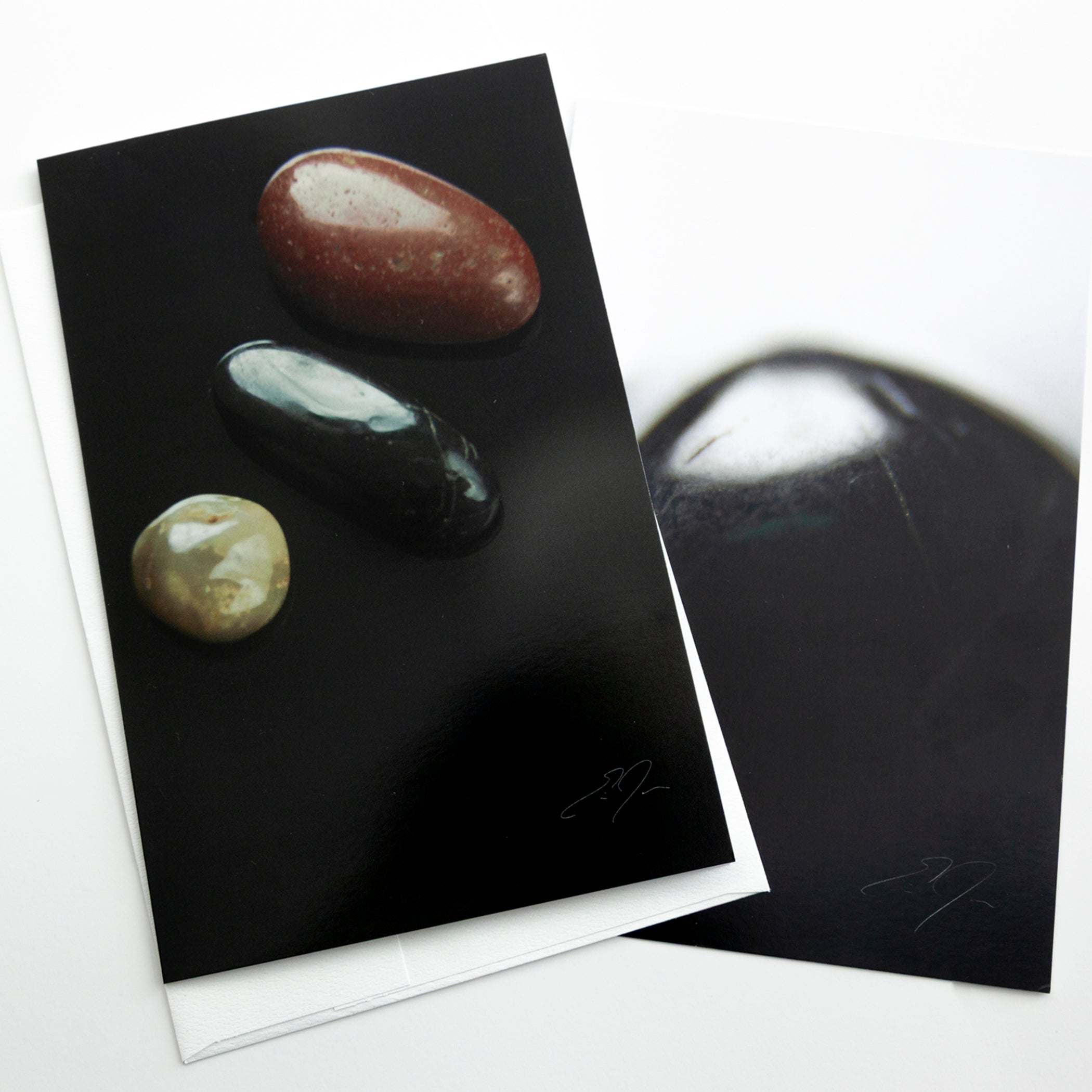 A square art print or card featuring three polished stones on a black background, shown overlapping another copy of the same print on a white surface.
