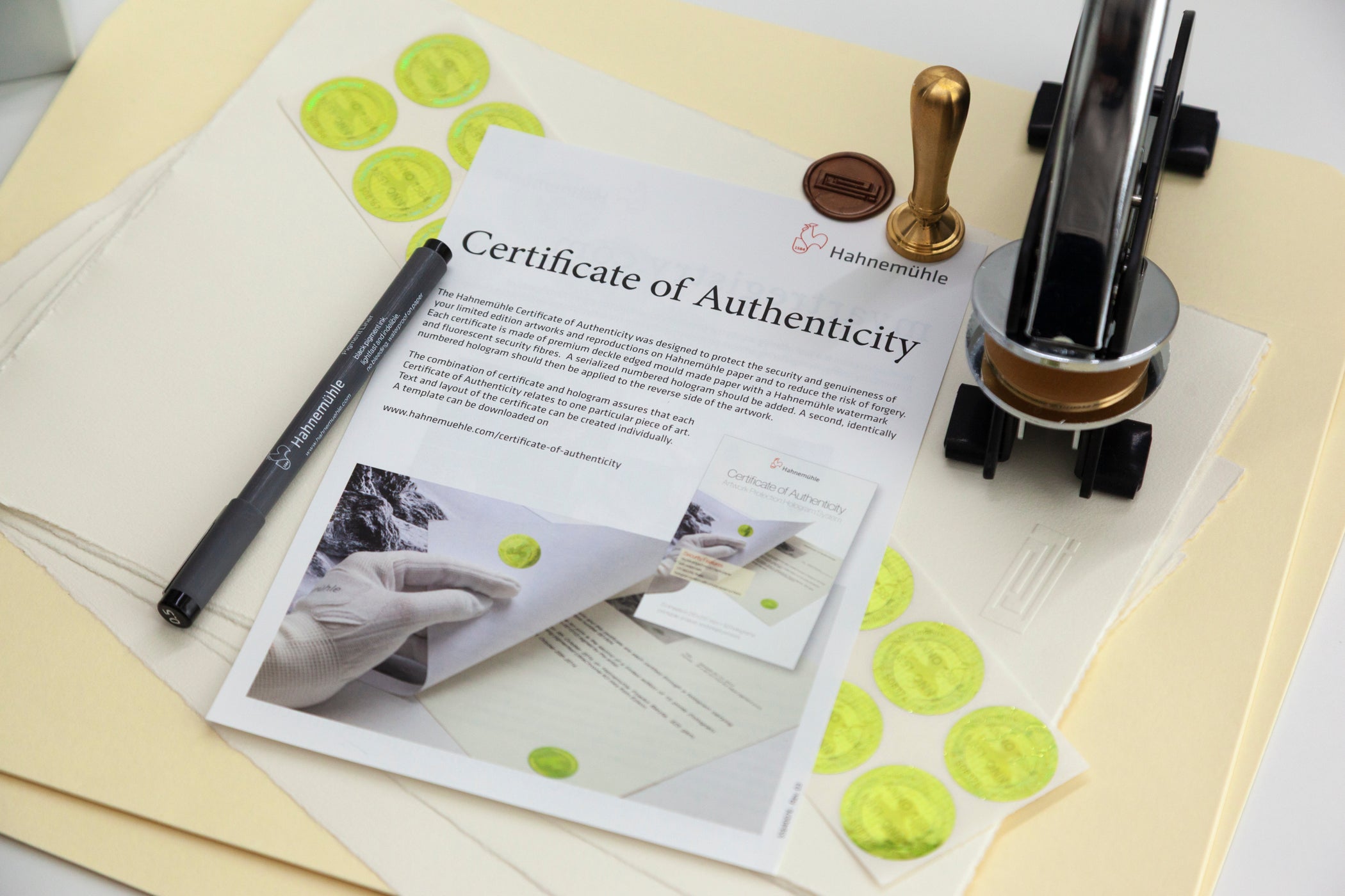 A flat-lay of authenticity documents and sealing tools, featuring a Certificate of Authenticity printed on paper, several round green seals, a black pen, a wooden stamp handle, and a clear acrylic stamp press on a light beige background.