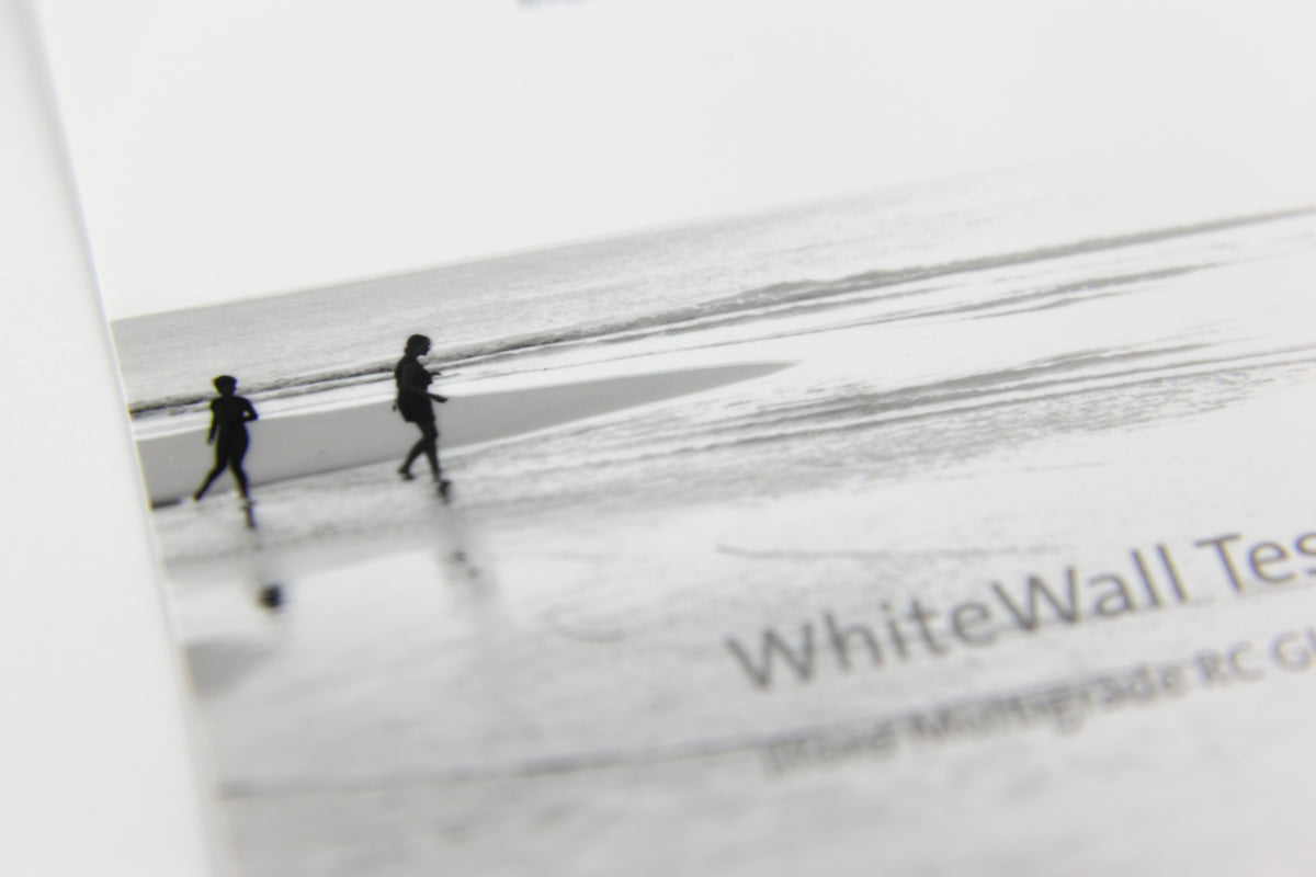 A close-up of a printed page featuring a black-and-white beach scene with two small silhouetted figures walking along the shoreline. Partial visible text reads 'WhiteWall Test.