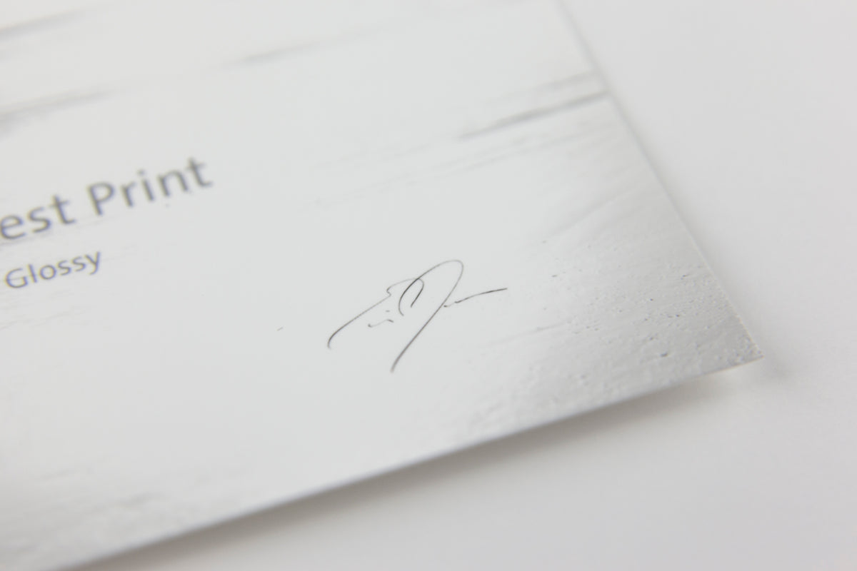 Close-up of a glossy white print card or paper sample with partial text reading 'est Print' and 'Glossy', plus a small printed signature, on a white background.